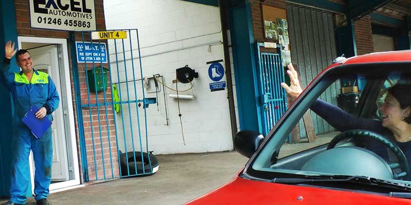 happy customer waving at an excel mechanic and driving away from excels garage in her little red car